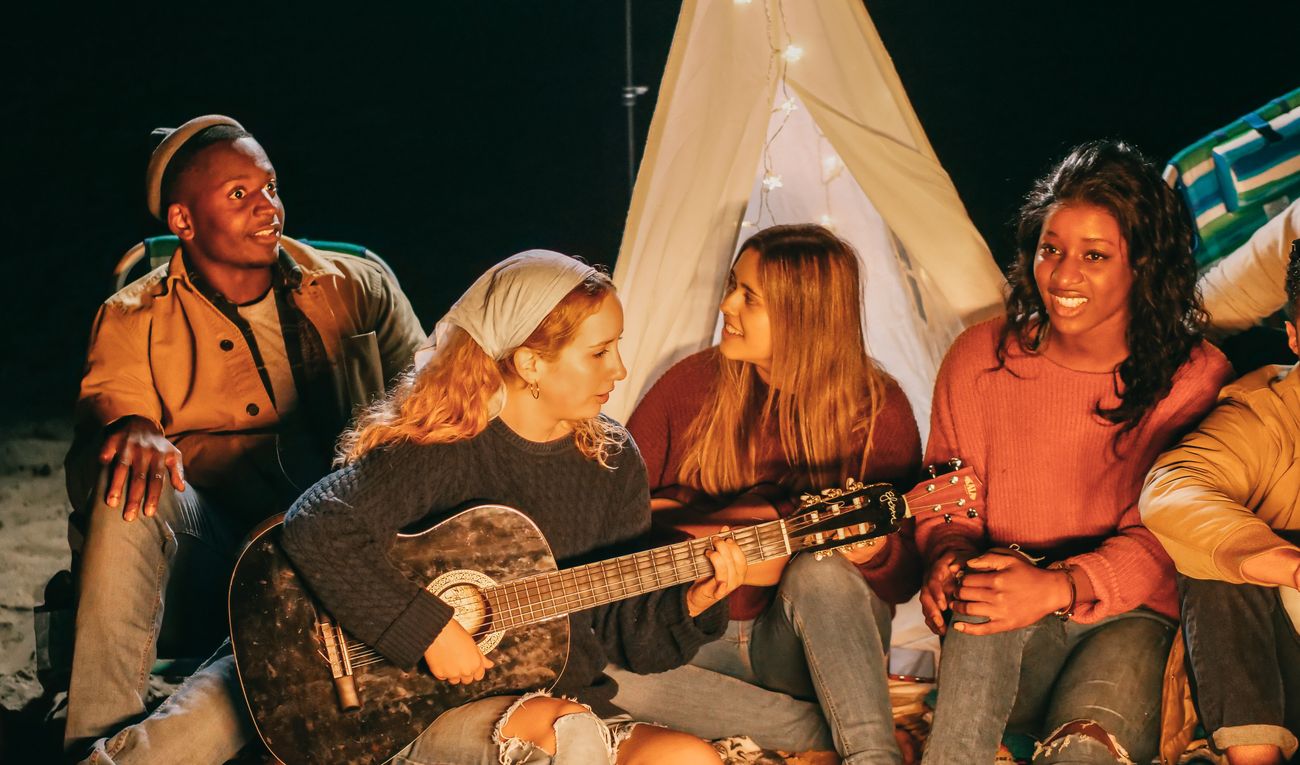 A diverse group of friends laughing together around a campfire, one playing guitar