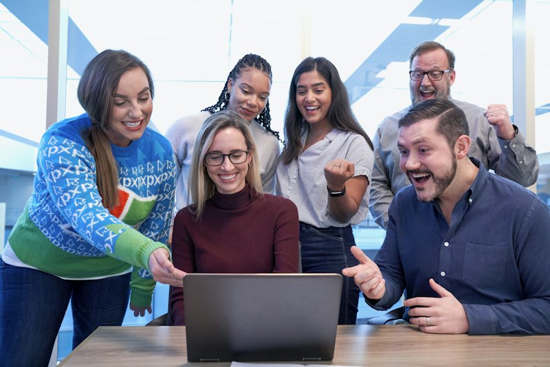 A diverse group gathered around a laptop, smiling and cheering
